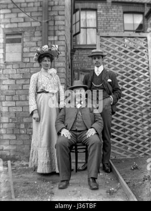 Edwardian family of three pose for a portrait in the backyard of a house in 1910. A bearded gentleman sits in a chair with a lady and a young man at his side. The younger man is wearing a straw boater hat. Restored using a high resolution scan taken from the original negative. Stock Photo