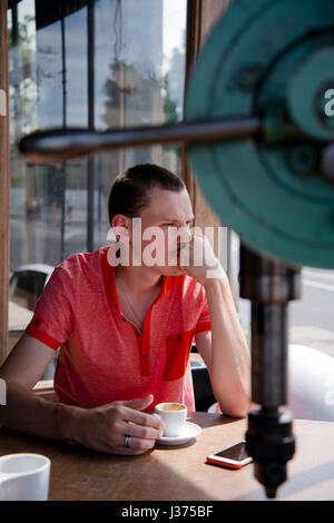 A sensitive young man with short hair looking thoughtfully out of the window while sitting at a table with a cup of coffee in hand, sadly discussing t Stock Photo