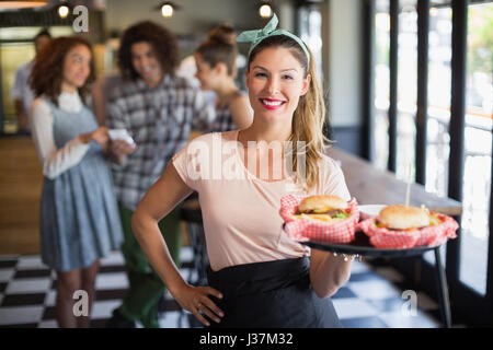 Portrait of smiling young waitress serving burger with customers in background at restaurant Stock Photo