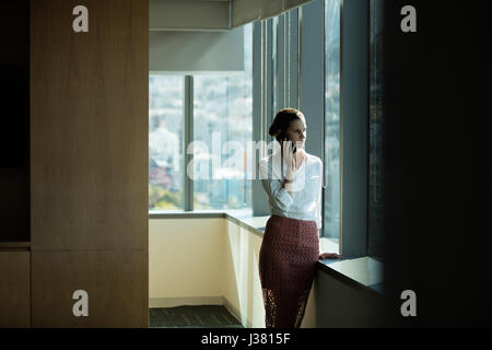 Businesswoman talking on phone while standing by window in office Stock Photo