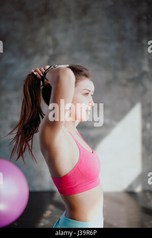 Womans break in workout to tie her hair. Female athlete tying hair before her workout with dumbbells. Beautiful brunette sitting at gym with red towel Stock Photo