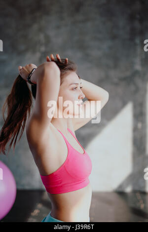 Womans break in workout to tie her hair. Female athlete tying hair before her workout with dumbbells. Beautiful brunette sitting at gym with red towel Stock Photo