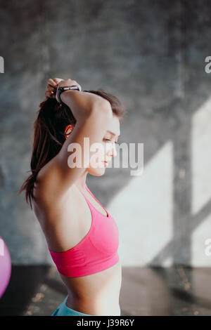 Womans break in workout to tie her hair. Female athlete tying hair before her workout with dumbbells. Beautiful brunette sitting at gym with red towel Stock Photo