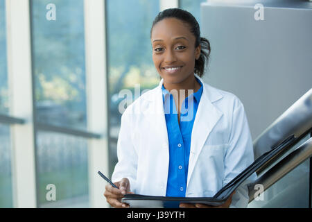 Closeup headshot portrait of friendly, smiling confident female doctor, healthcare professional with labcoat, holding pen to face and holding notebook Stock Photo