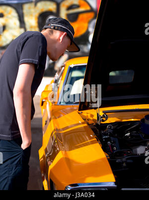 A young man in a cap with hands in pockets examines classic vintage muscle bright yellow car with open hood standing in the street on the day of parad Stock Photo