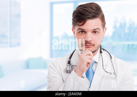 Portrait of handsome pensive doctor in white coat keeping hand on his chin and thinking with copy space Stock Photo