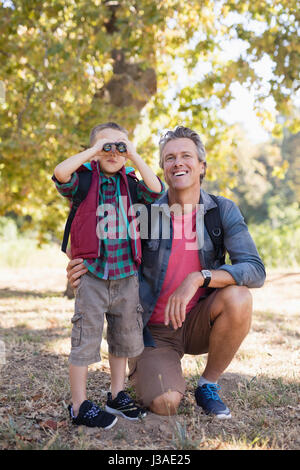 Happy mature father kneeling by boy looking through binoculars in forest Stock Photo