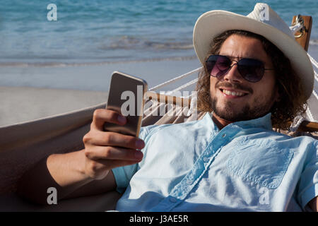 Smiling man relaxing on hammock and using mobile phone on the beach Stock Photo
