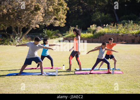 Young female trainer watching children praciticing Virabhadrasana II pose on mat Stock Photo