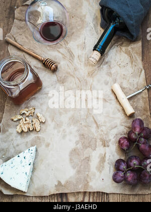 Wine and appetizer set with copy space in center. Glass of red wine, bottle, corkscrewer, blue cheese, grapes, honey, walnuts on oily craft paper over rustic wooden table, top view. Stock Photo