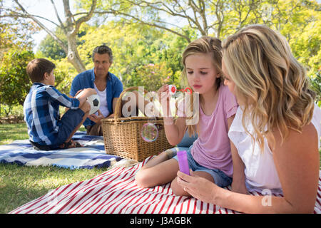 Mother and daughter blowing bubble while father and son playing with football in park Stock Photo