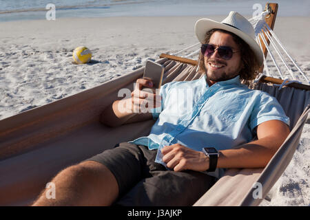 Smiling man relaxing on hammock and using mobile phone on the beach Stock Photo