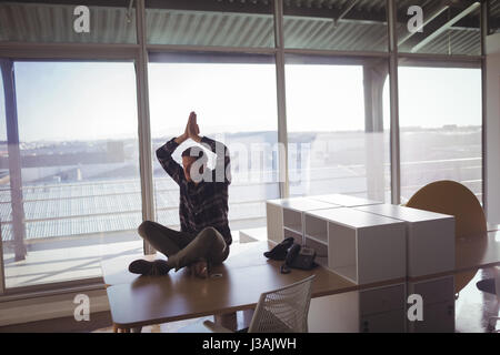 Young businessman meditating on desk against glass window at office Stock Photo
