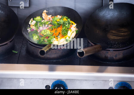 Sliced vegetables from cabbage, broccoli, carrots, onions and zucchini stew in a large sooty industrial frying pan on three-plank stove - healthy food Stock Photo