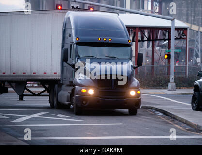 A large classic dark big rig semi-truck with a turn on headlights and dry van trailer for the transportation of commercial goods over long distances Stock Photo