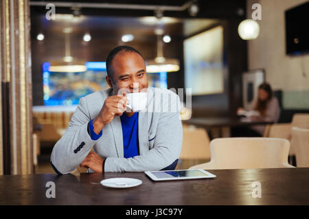 Handsome Afro American man is using a tablet, holding a cup and smiling while sitting near the window at home Stock Photo