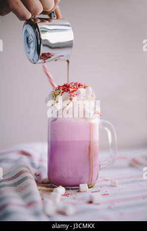 Woman is pouring coffee in cup of colored milk with cream, marshmallow and colorful decoration. Milk shake, cocktaill, frappuccino.  Unicorn coffee, u Stock Photo