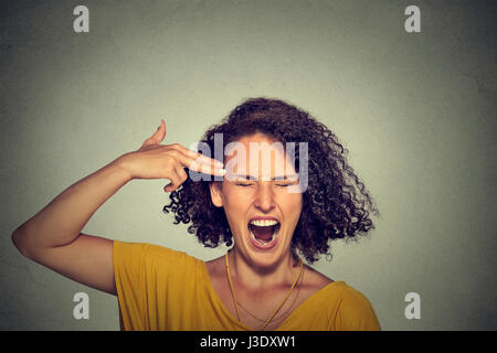 Headshot portrait young woman committing suicide with finger gun gesture isolated on grey wall background. Human emotions face expressions. Overworked Stock Photo