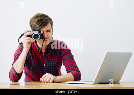 Young and successful man uses Mirrorless camera and sits at a table with a laptop in the office Stock Photo