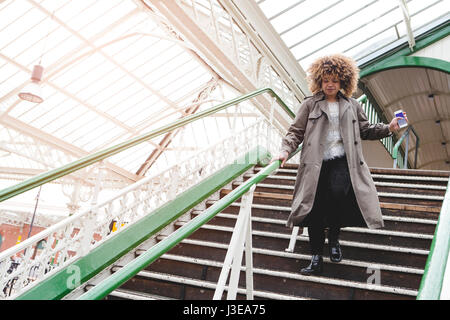 Woman running down the stairs at a train station with her phone in hand. She is late for her train. Stock Photo