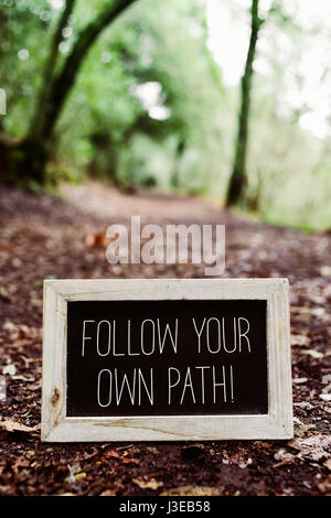 closeup of a wooden-framed chalkboard with the text follow your own path written in it, placed on the ground in a path in the forest Stock Photo