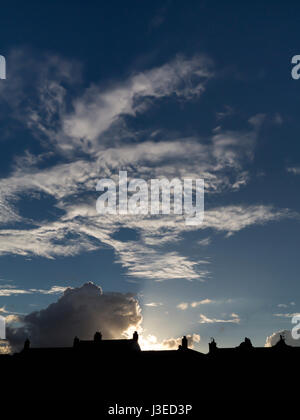 Sun setting behind silhouetted rooftops with fanning rays in to a dark blue mottled sky Stock Photo