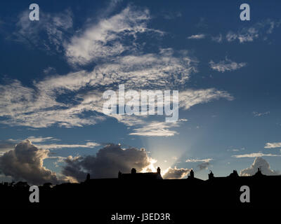 Sun setting behind silhouetted rooftops with fanning rays in to a dark blue mottled sky Stock Photo