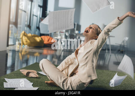 Excited young businesswoman throwing documents while sitting on floor in office Stock Photo