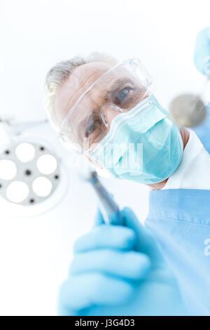 Professional mature dentist in medical mask and protective glasses looking at camera Stock Photo