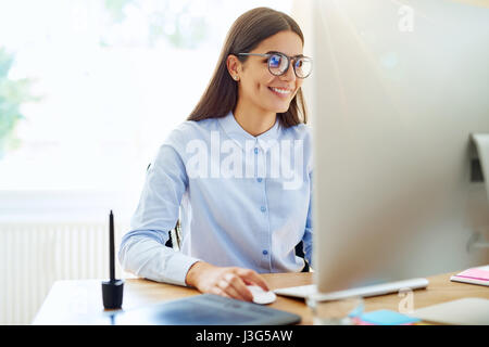 Smiling female graphic designer using mouse, stylus and tablet next to her on wooden desk in small office Stock Photo