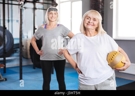 Lets play. Happy grey haired senior woman having a ball in her hands and standing in front of her friend while visiting the sport centre Stock Photo