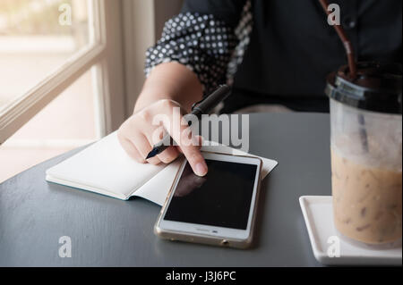 Woman hand touching on smartphone screen while writing something in coffee shop. Working from anywhere concept Stock Photo