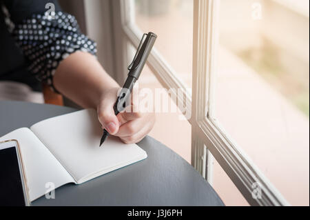 Left-handed woman holding pen while writing on small notebook beside window. Freelance journalist working at home concept. Stock Photo