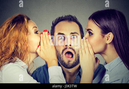 Two young women telling whispering secret gossip in the ear to an amazed shocked man with wide open mouth. Human emotions face expression reaction. Ho Stock Photo