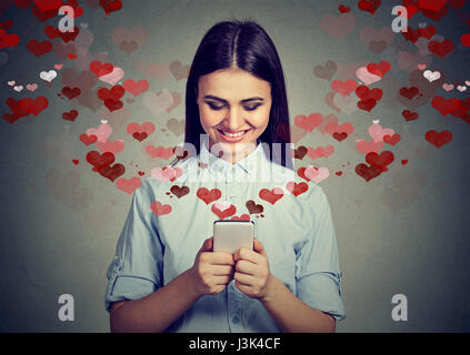 Beautiful happy woman sending love text message on mobile phone with red hearts flying away from screen isolated on gray background. Positive human em