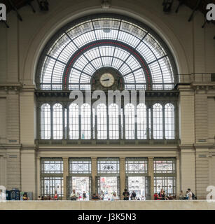 Many people wait inside the front doors of the Budapest Keleti Railway Station, in front of the big decorative windows. Stock Photo