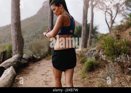 Female runner checking fitness progress on her smart watch. Asian woman using fitness app to monitor workout performance, while walking through mounta Stock Photo