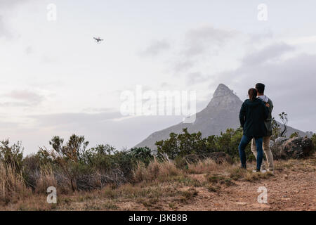 Young couple standing together outdoors watching and navigating a flying drone in sky over countryside. Man and woman using drone for photography in r Stock Photo