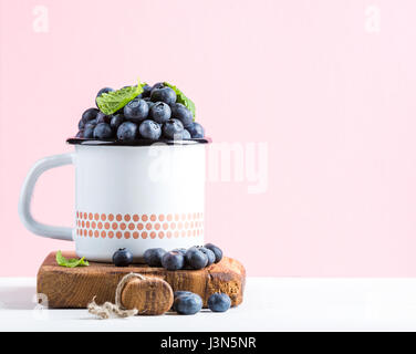 Fresh ripe blueberries in country style enamel mug on rustic wooden board over pastel pink background Stock Photo