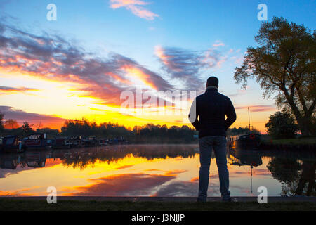 Rufford, Lancashire, 7th May 2017. UK Weather.  A man watches a beautiful sunrise & early morning mist as the sun tries to break through the clouds over the canal boats moored on Rufford Marina in Lancashire.  This local beauty spot is located just off the Leeds/Liverpool canal.  With it's stunning viewpoints and on site cafe's, it is a very popular resting place for those travelling this famous East to West route.  Credit: Cernan Elias/Alamy Live News Stock Photo