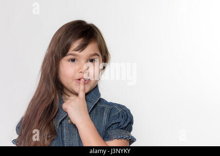 Portrait of slightly smiling brunette long haired little girl gesturing shush. All is on the gray background. Stock Photo