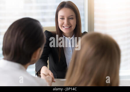 Attractive cheerful businesswoman and couple shaking hands over a desk closing real estate deal or partnership, finishing up or starting meeting, esta Stock Photo