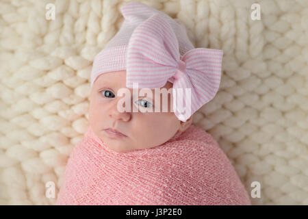 Studio portrait of an alert, month old, newborn baby girl. She is wearing a hat with a large bow and wrapped in a pink swaddle. Stock Photo