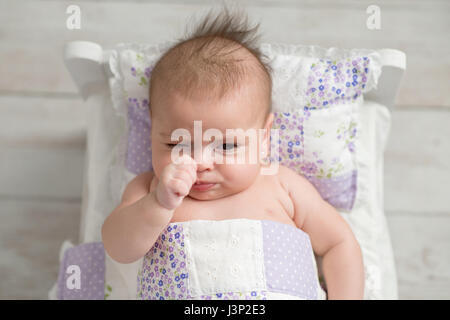 An overhead shot of a four month old, sulking, baby girl. She is lying in a tiny bed with quilted bedding. Stock Photo
