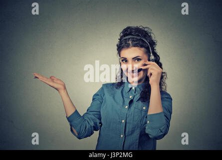 Closeup portrait of beautiful, adorable smiling female customer representative with phone headset pointing at copy space isolated on gray wall backgro Stock Photo