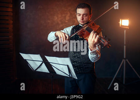 Portrait of male violinist with violin against music stand. Fiddler man with musical instrument playing in studio, solo concert training Stock Photo