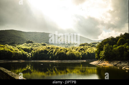 Lake in the middle of the forest with sunrays behind clouds Stock Photo