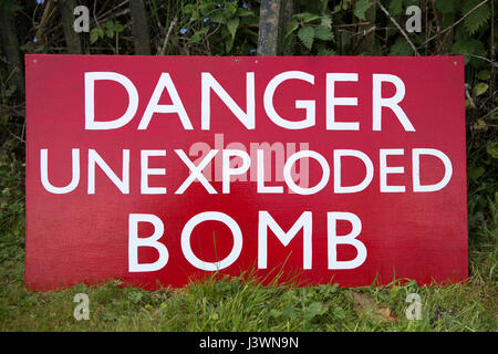 Red signboard with white writing saying DANGER UNEXPLODED BOMB. Sign resting on the grassy ground. Stock Photo