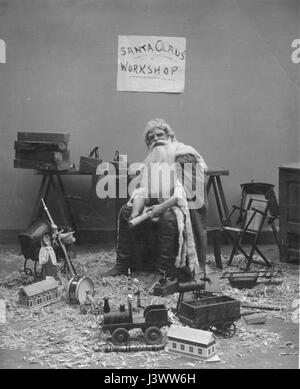 'Santa Claus' Workshop'  Santa is whittling a new toy, while sitting in his workshop, c. 1908.  Completed toys around him are a drum, simple train engine with car, dolls and a toy baby carriage, a small hobby horse, a two-hand whistle, other small toys and lots of wood shavings. His work table holds wood planes, wood to be carved, and an ax and a saw hang from the table's side.  To see my other Christmas-related vintage images, Search:  Prestor  vintage  holiday Stock Photo