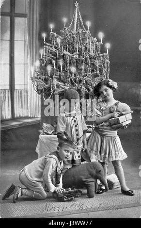 Two boys and a girl hold gifts - toy Elephant, toy rabbit, train engine, toy whip and other gifts wrapped in plain brown paper - as they stand in front of a Christmas tree decorated with lighted candles. 1915.  To see my other Christmas-related vintage images, Search:  Prestor  vintage  holiday Stock Photo